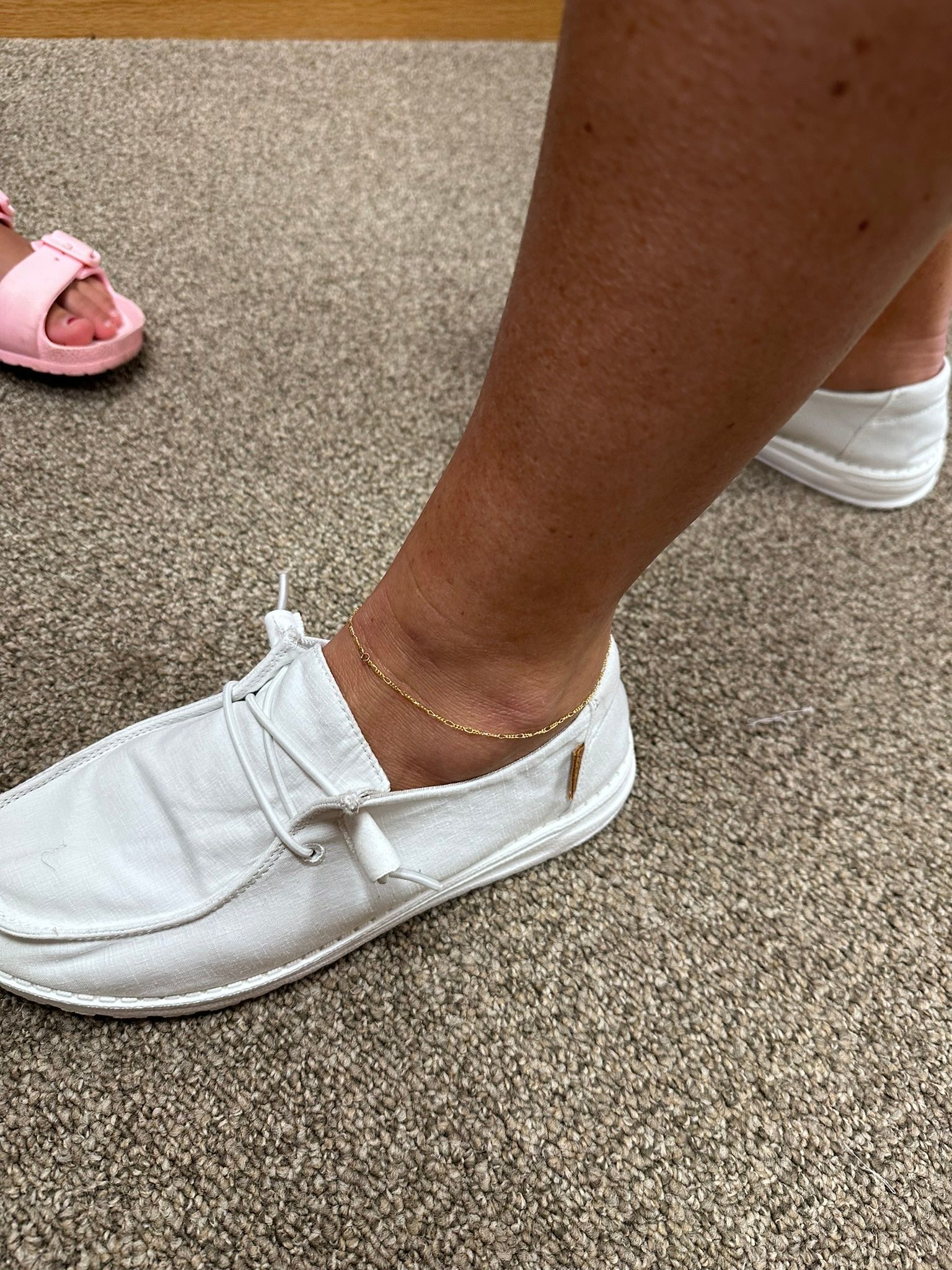 Person's ankle with a gold anklet and white shoe on a carpet. A child's pink sandal is partially visible.