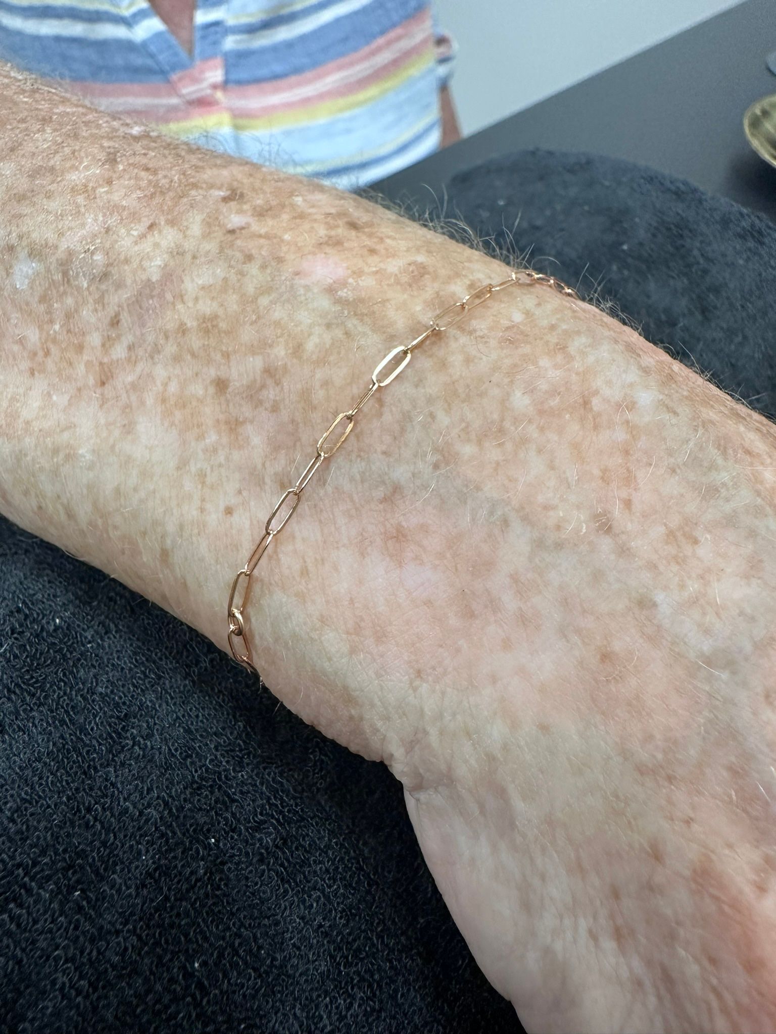 Arm with delicate gold bracelet, freckled skin, resting on black surface.