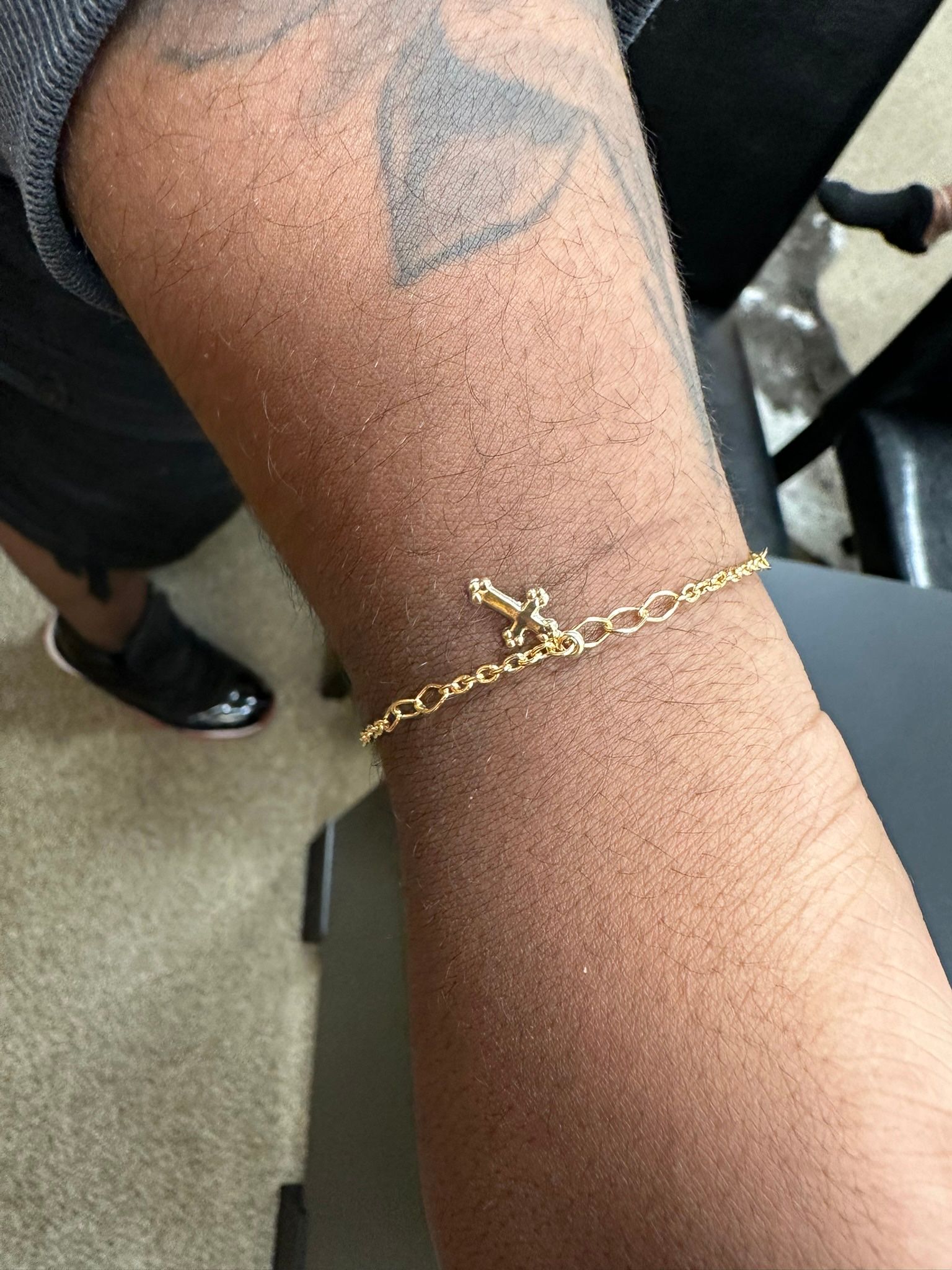 Close-up of a person's arm with a gold chain bracelet featuring a decorative clasp. Dark skin, tattoo visible.