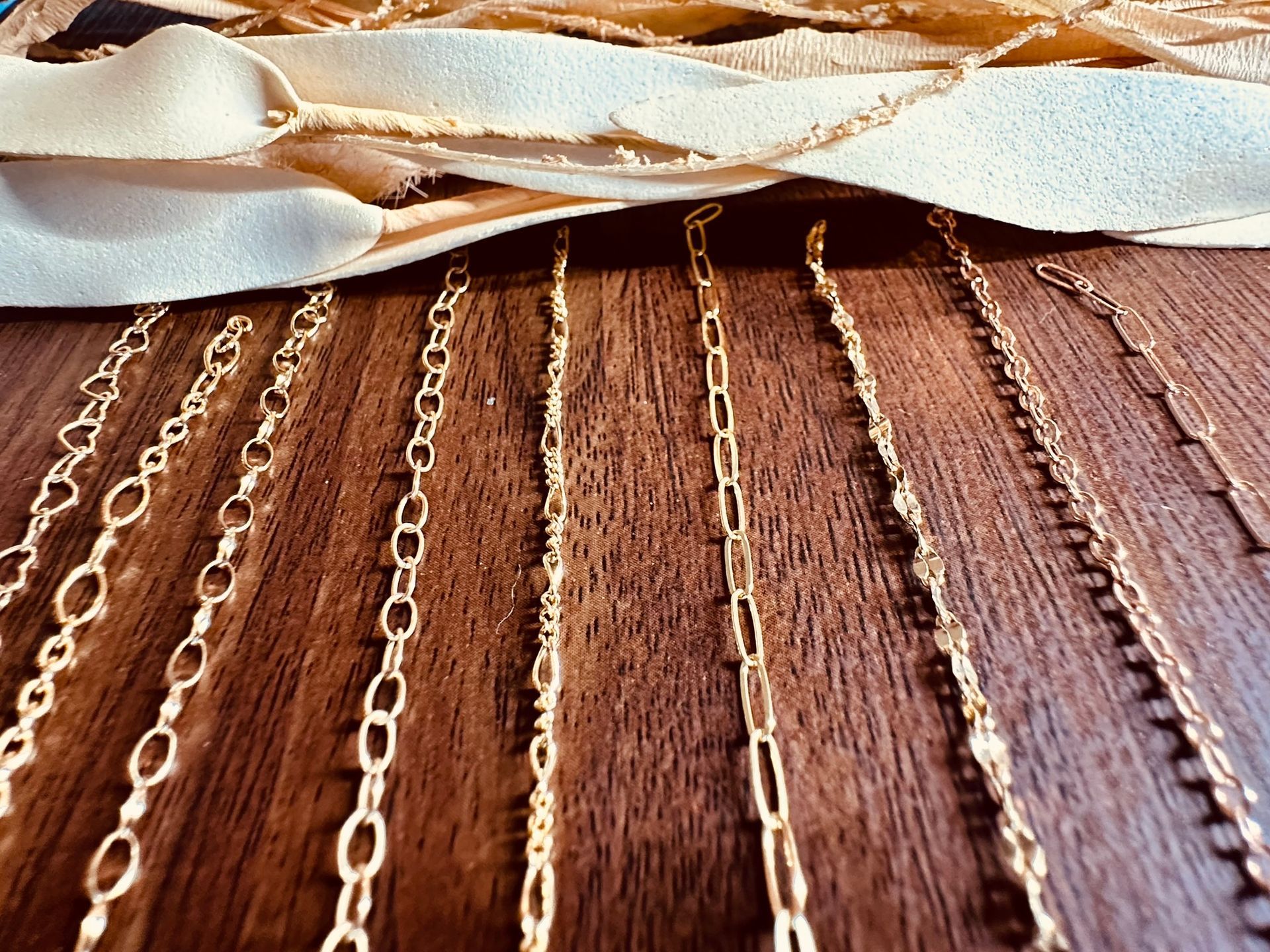 Close-up of gold chain necklaces on a wooden surface, with a cream-colored fabric strip in the background.