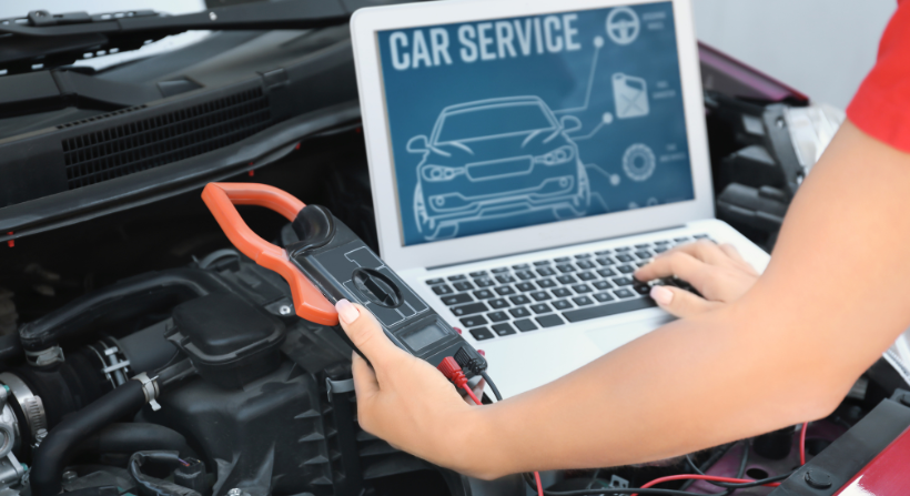 A person holding a clamp meter and using a laptop to run car diagnostic software while working on an open engine bay.