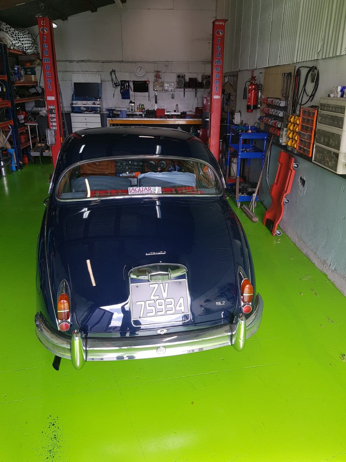 Dark blue vintage sedan parked in a garage with a green floor, between two red automotive lift posts.