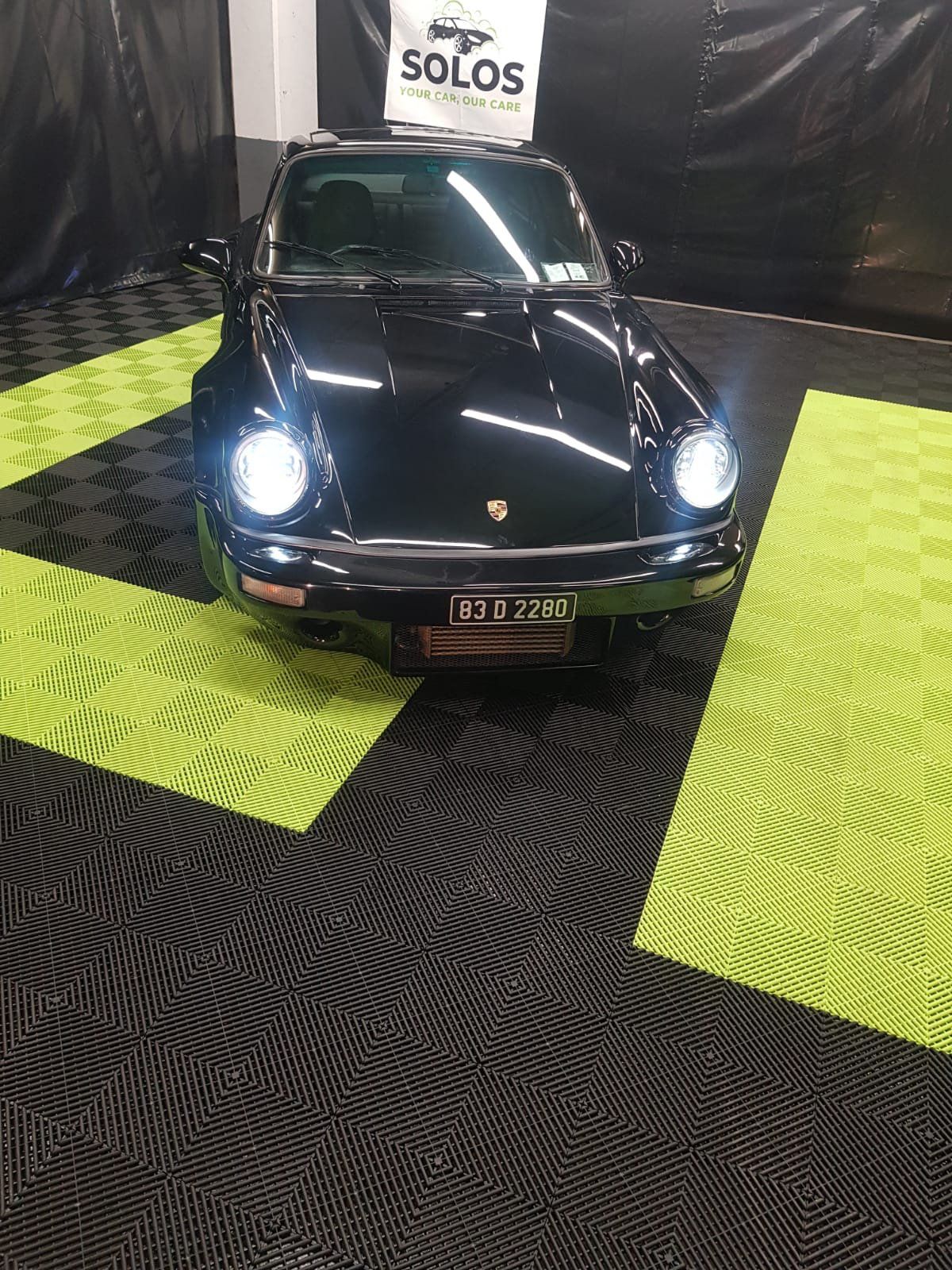 A black classic Porsche 911 parked on a black and green interlocking tile floor, with a