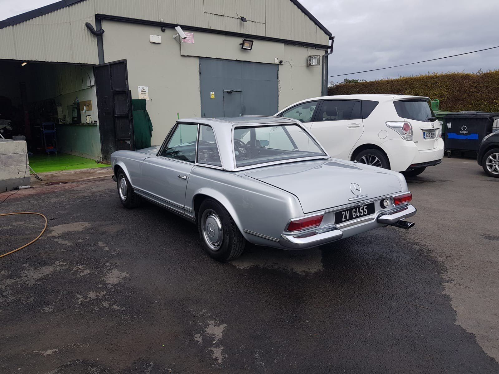 A silver vintage Mercedes-Benz convertible parked outside an industrial building next to a modern white car.