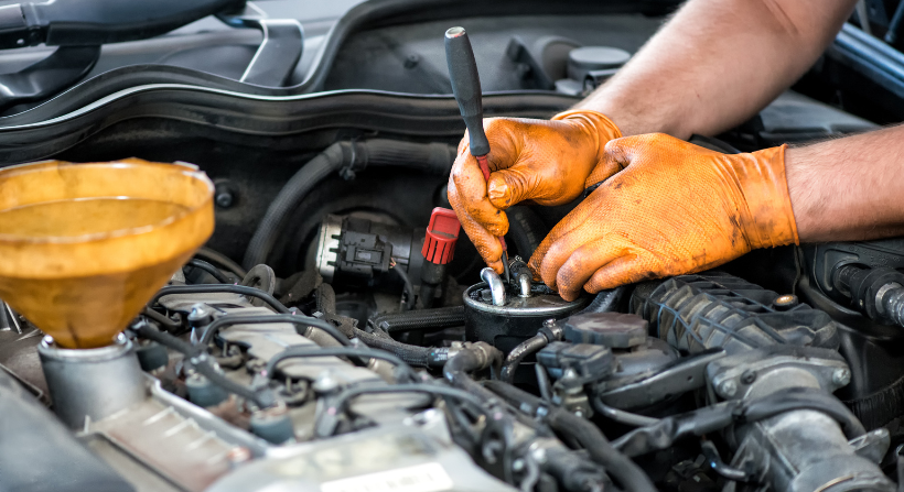 A person wearing orange gloves uses a screwdriver to work on a car engine with a yellow funnel nearby.