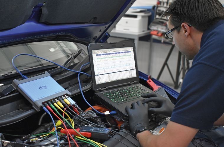 A technician wearing black gloves uses a laptop and diagnostic equipment to test a car engine in an auto repair shop.