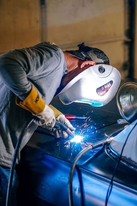 A person wearing protective gear and a welding helmet uses a torch to weld a metal surface, creating bright sparks.