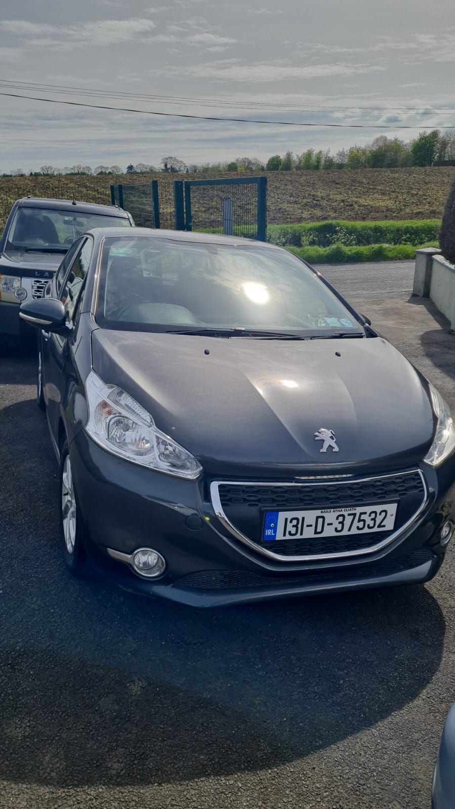 A dark grey Peugeot 208 parked on a paved lot with a sunny, rural background.