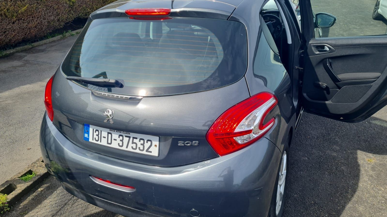 A gray Peugeot 208 parked outdoors, viewed from the rear with the passenger door open.