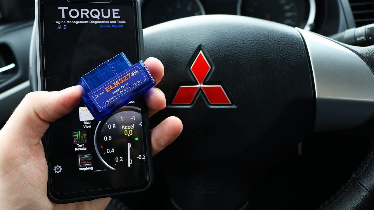 A hand holds a blue OBD2 scanner against a phone running the Torque app, with a Mitsubishi steering wheel in the background.