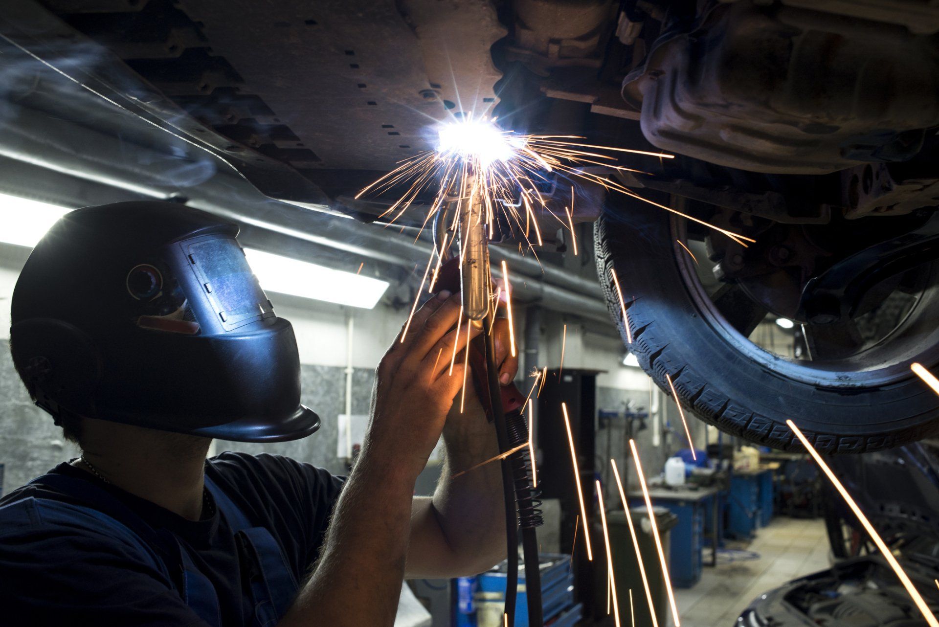 A welder wearing a protective mask works on the underside of a car, creating bright sparks in a garage workshop.