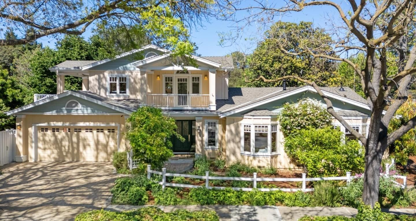 A two-story light yellow house with a two-car garage, a balcony, and a white picket fence, surrounded by green trees.