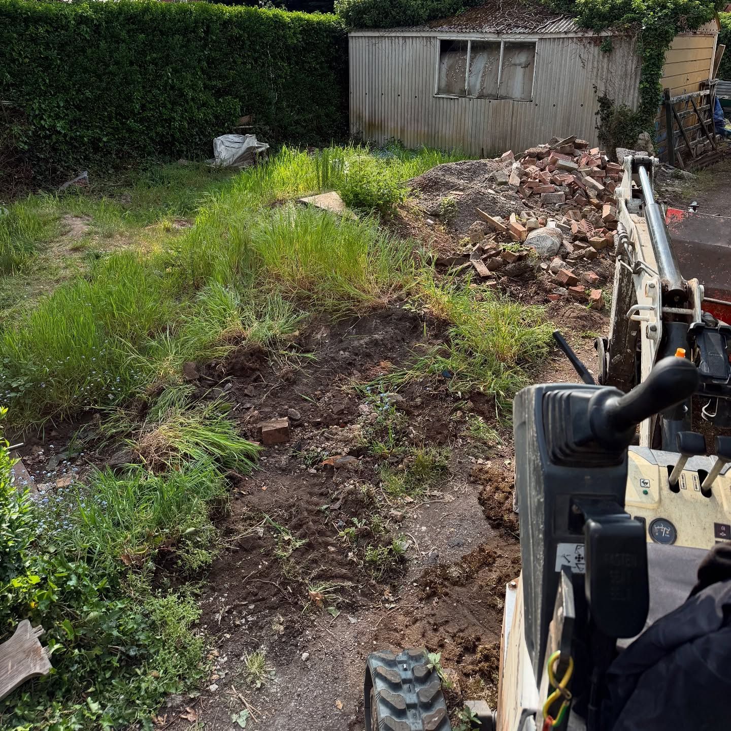 Backyard with overgrown grass, dirt pile, and rubble beside a small shed, viewed from a digger.