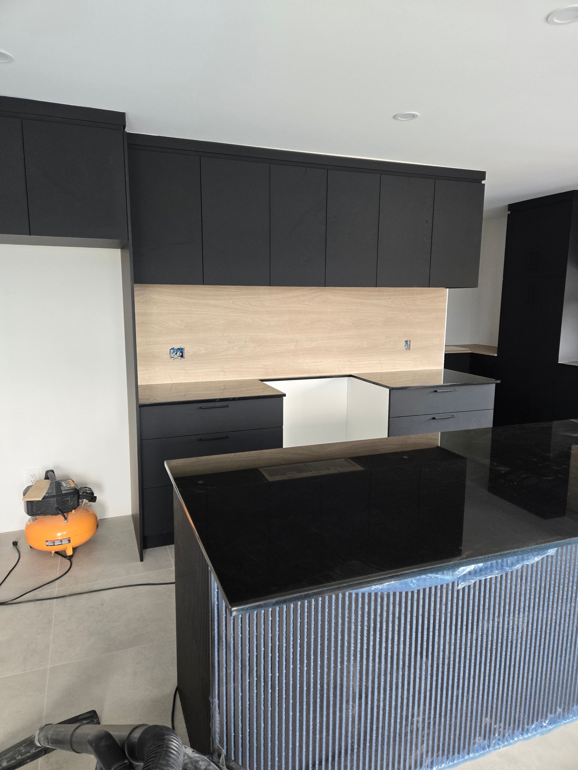Cuisine moderne en construction avec armoires noires, îlot et crédence en bois clair.