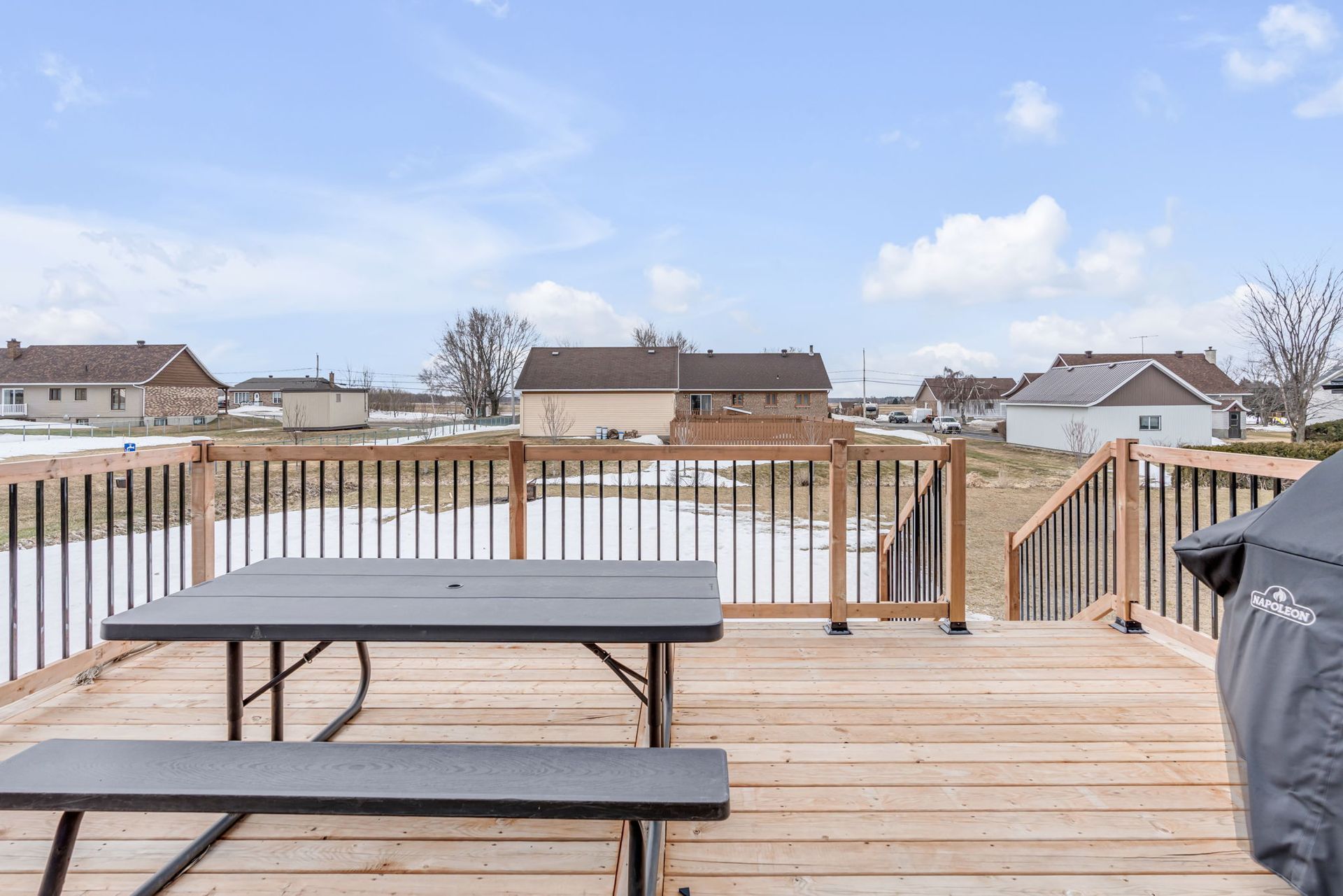 Terrasse en bois avec table de pique-nique et barbecue noir, donnant sur des jardins et des maisons enneigés sous un ciel nuageux.