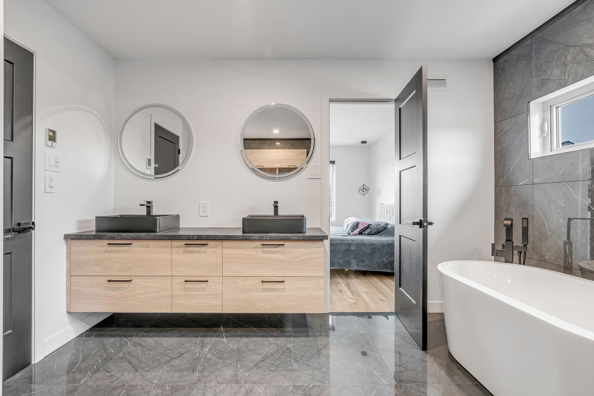 Salle de bains moderne avec meuble-lavabo suspendu en bois, deux lavabos noirs, grands miroirs, baignoire îlot et carrelage gris foncé au sol.