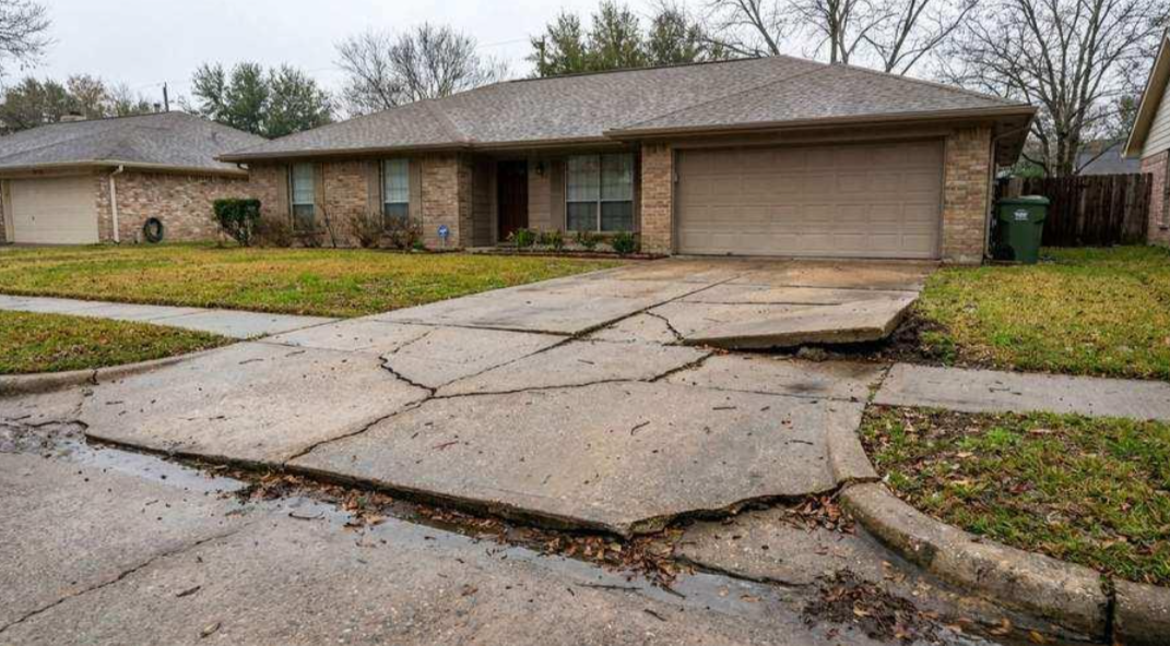 Cracked residential driveway leading to a brick house with a garage on a cloudy day