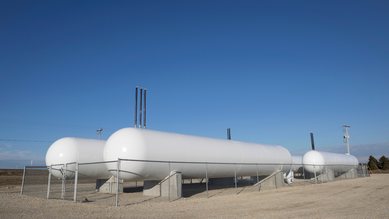 Three white cylindrical storage tanks behind a chain link fence, set against a clear blue sky.