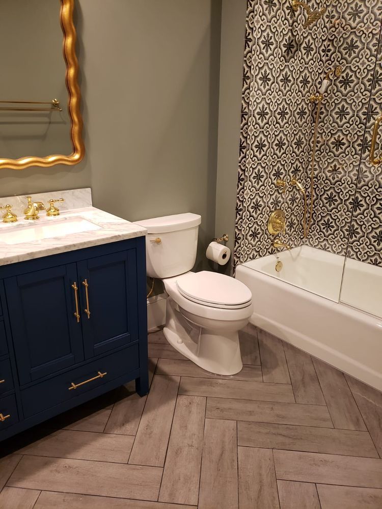 Bathroom with navy blue vanity, white toilet, patterned shower wall, gold fixtures, and gray herringbone floor.