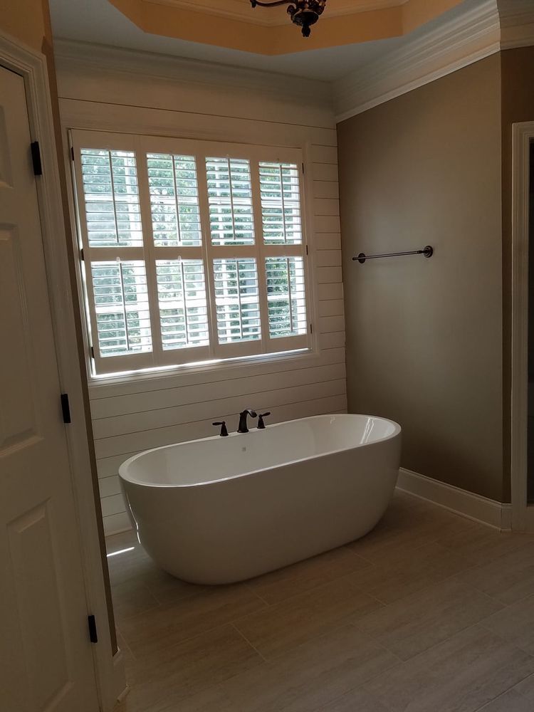 Bathroom with white tub, shutters, beige walls, and black fixtures.