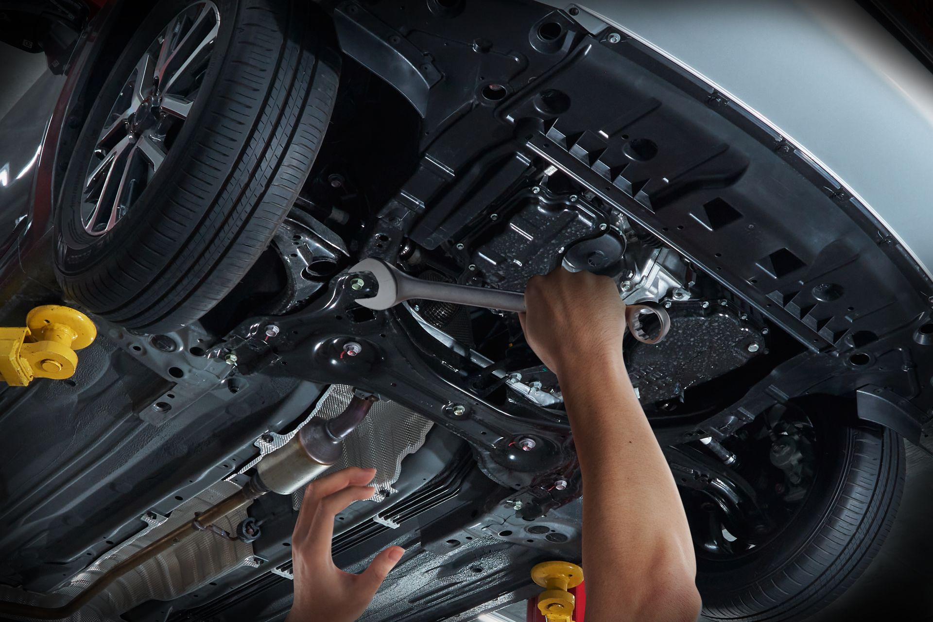 Mechanic working under a car on a lift, using tools near the suspension and exhaust.