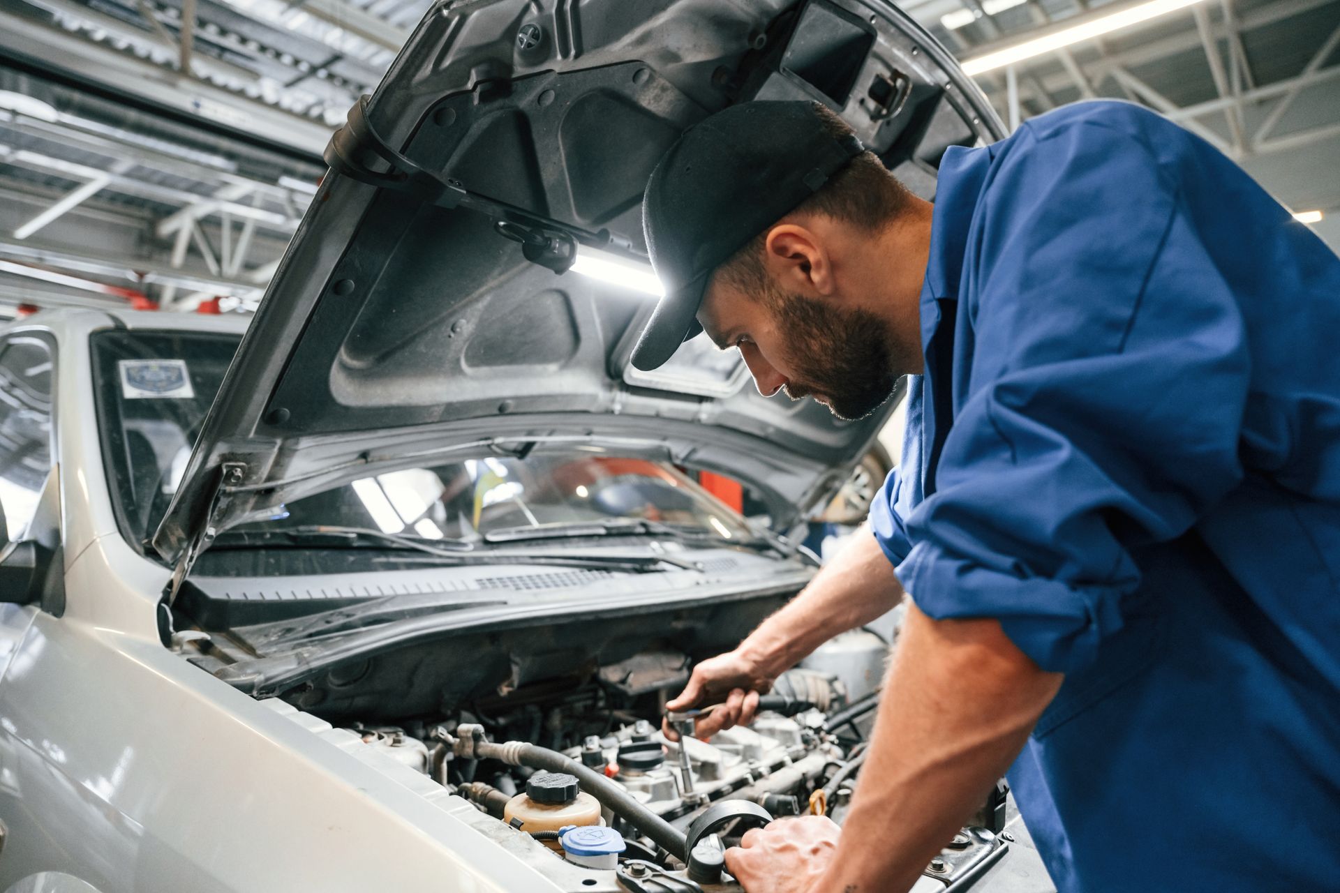 Mechanic in blue coveralls inspecting a car engine with the hood open in a workshop