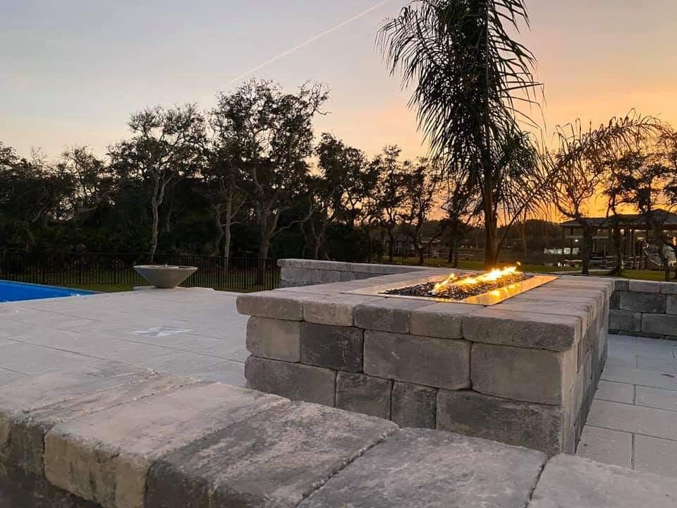 Stone fire pit glowing at sunset on a paved patio with trees in the background