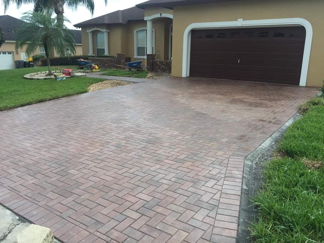 Brick driveway leading to a beige house with a dark garage door and green lawn