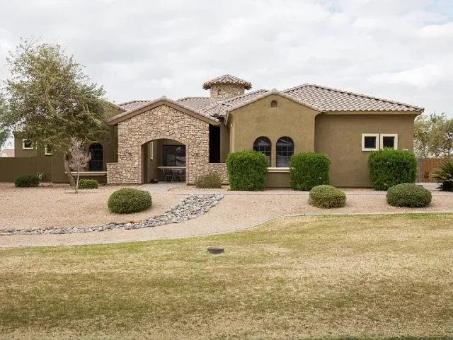 Single-story stucco house with tile roof, desert landscaping, and a curved driveway under a cloudy sky