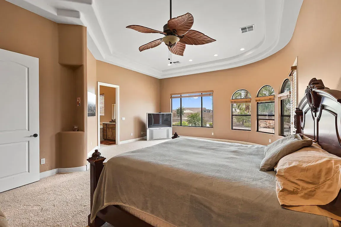 Spacious beige bedroom with a canopy bed, ceiling fan, and large windows overlooking a bright view