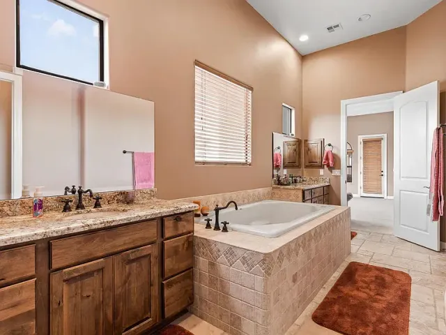 Spacious beige bathroom with double vanity, soaking tub, window blinds, and rust-colored rug