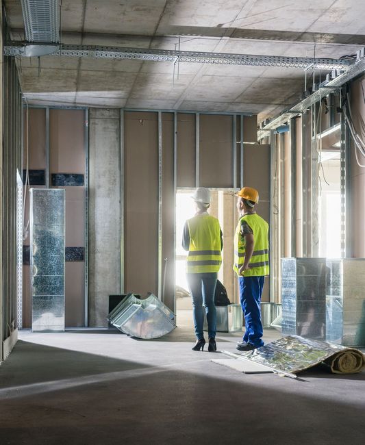 Two construction workers in safety vests and hard hats inspect an unfinished room under renovation.