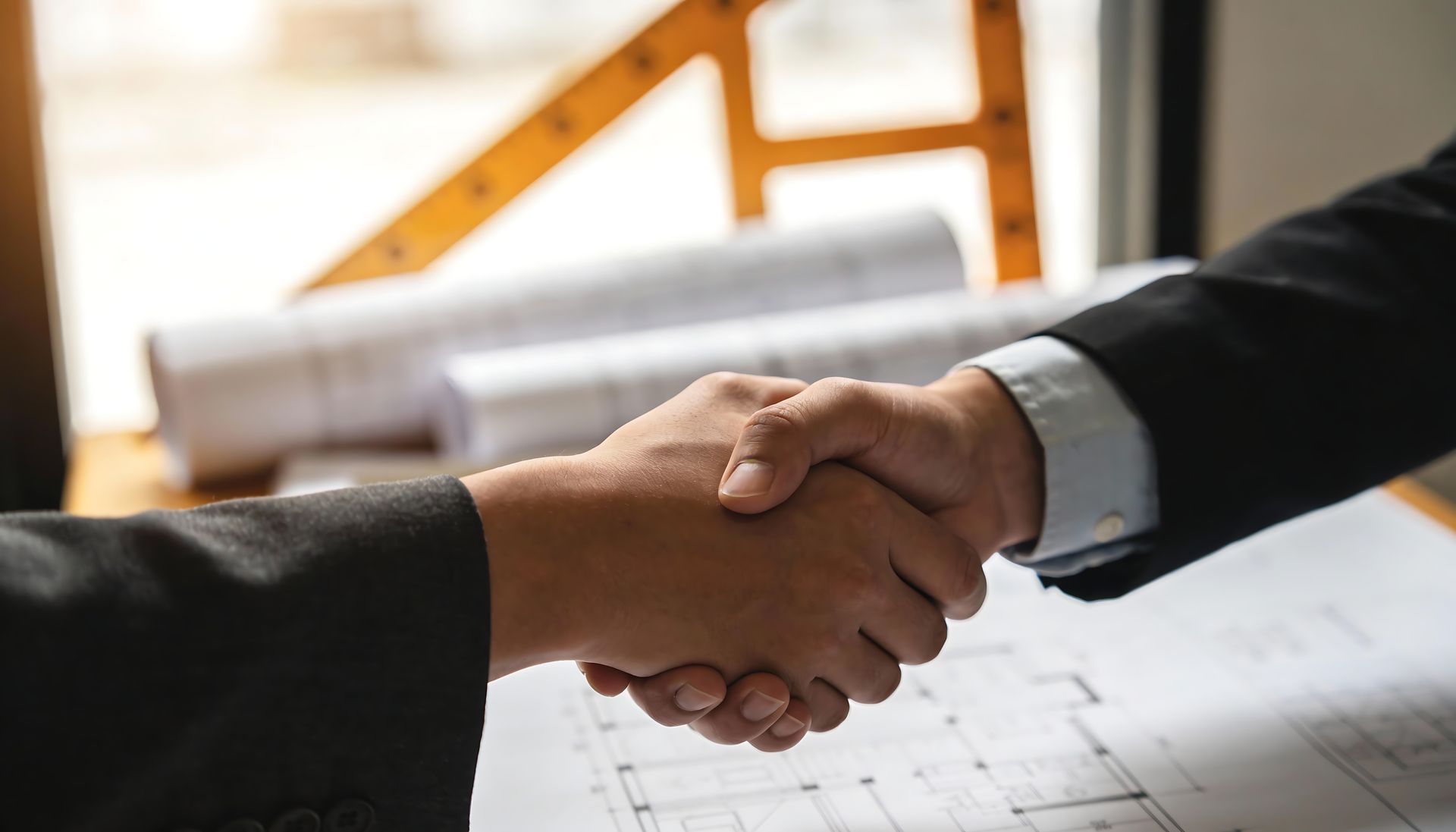 Two people in business suits shaking hands over a desk in an office.