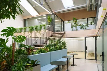 Bright atrium lounge with stairs, glass walls, plants, and blue seating under skylights