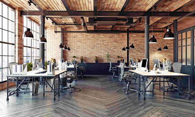 Open-plan office with exposed brick, wood beams, desks, chairs, and hanging pendant lights.