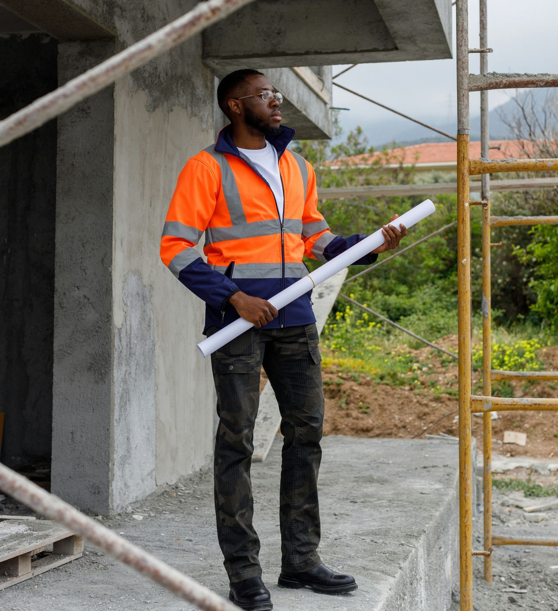 A man in an orange jacket is holding a blueprint in front of a scaffolding.
