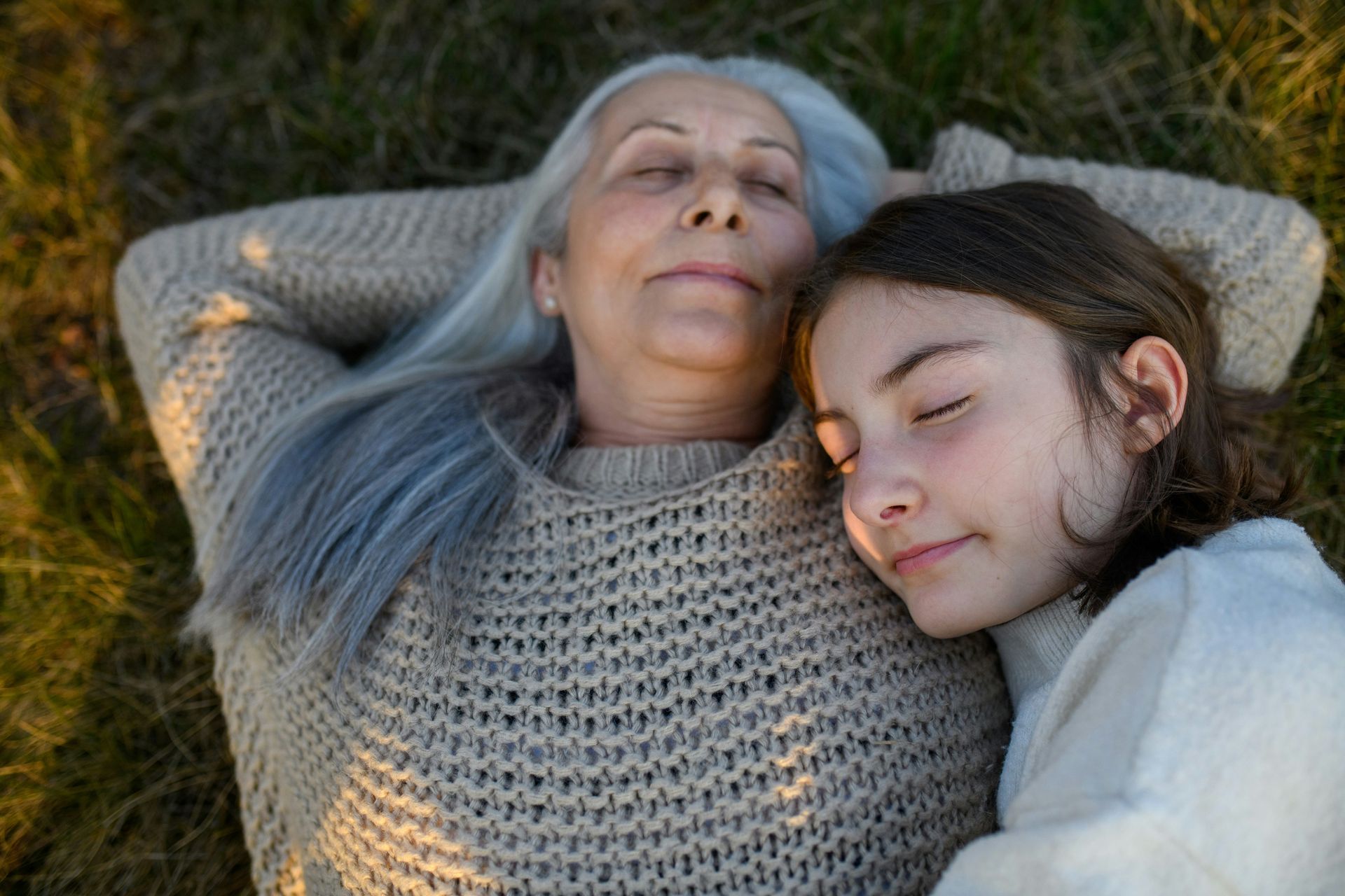 An elderly woman and a young girl are laying in the grass with their eyes closed.