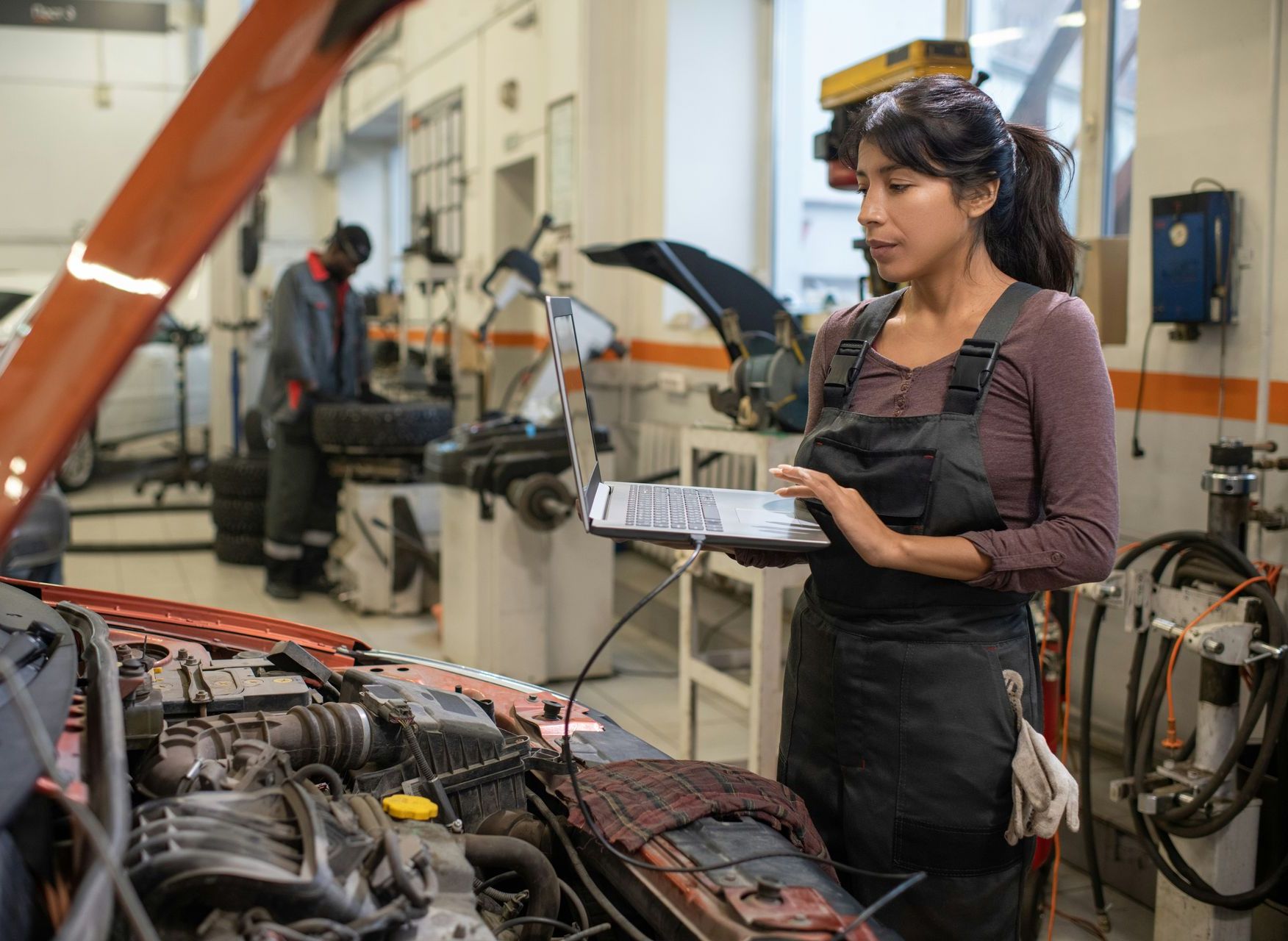 A woman is working on a car in a garage while holding a tablet.