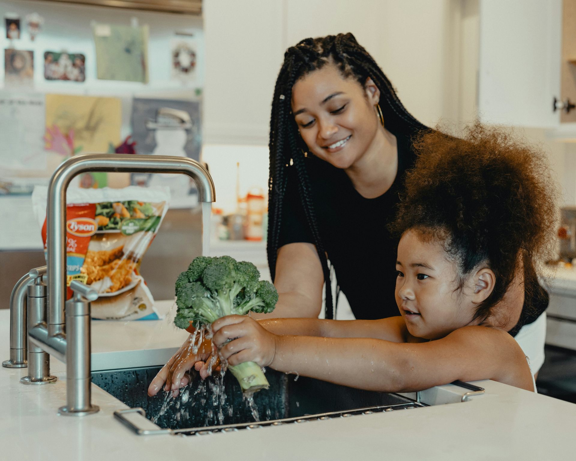 A woman and a little girl are washing broccoli in a kitchen sink.