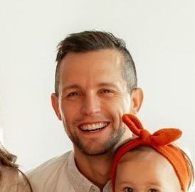 A man is smiling while holding a baby in an orange headband.