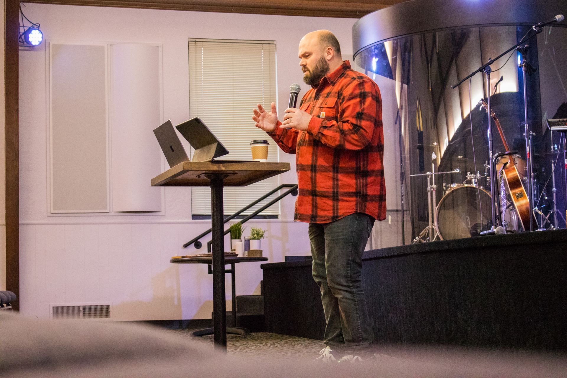 A man in a red plaid shirt is standing on a stage holding a microphone.