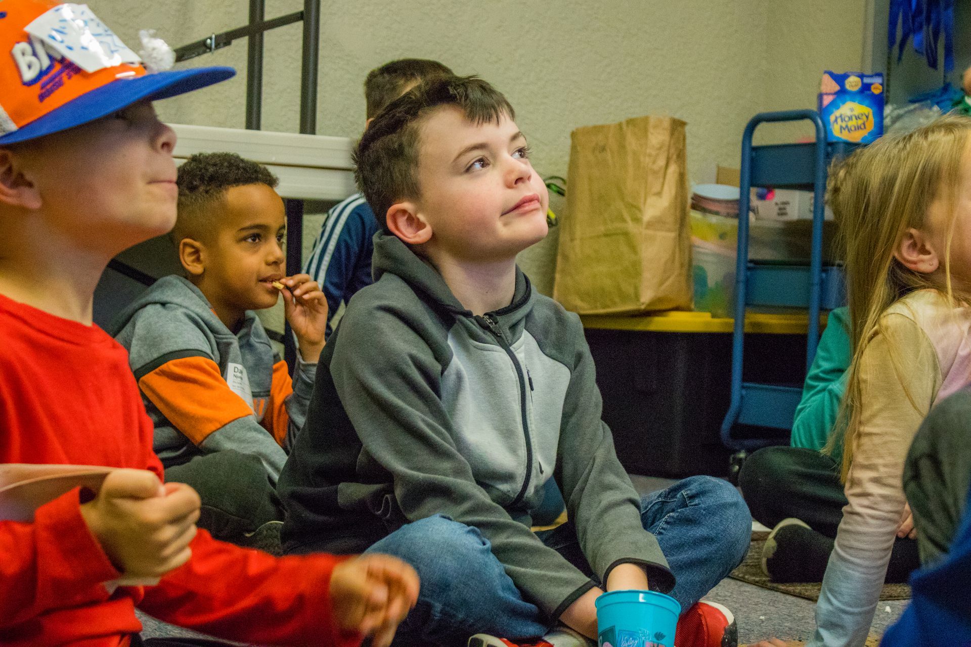 A group of children are sitting on the floor in a classroom looking up.
