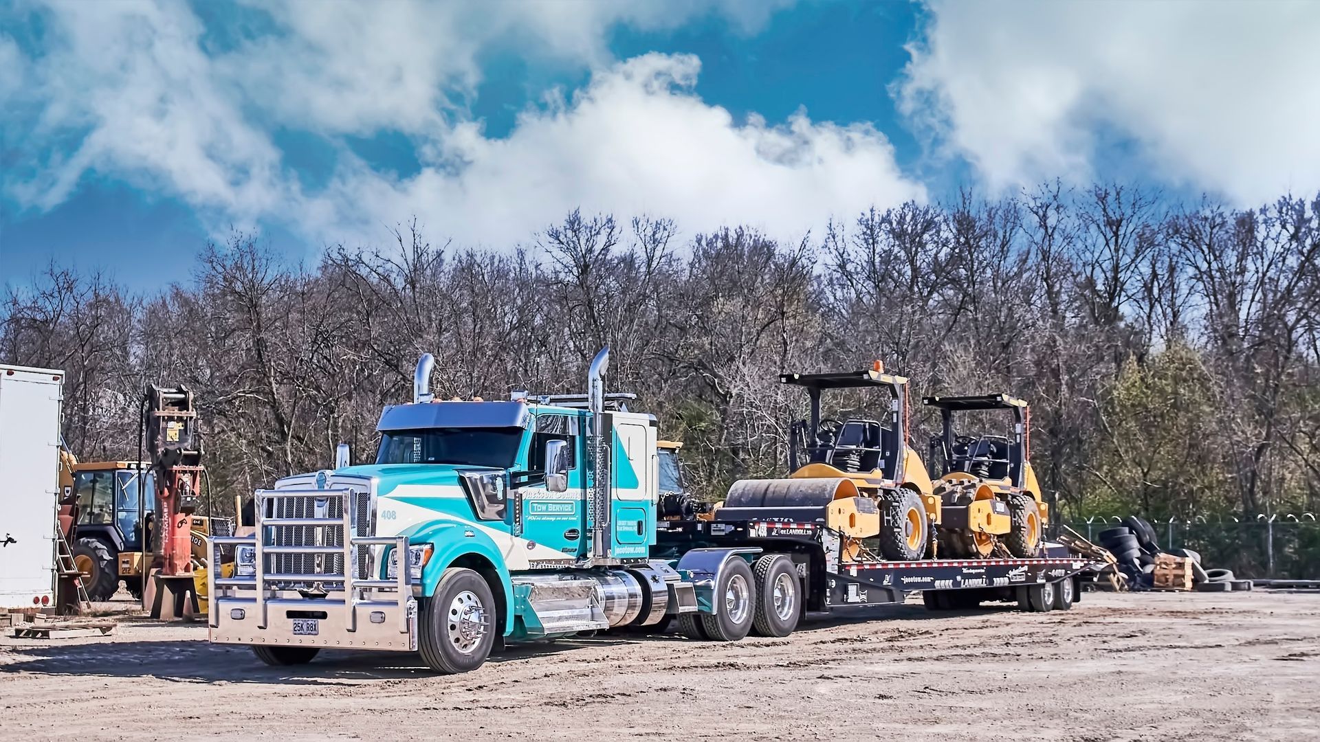 Tow truck hauling a blue semi and two yellow construction vehicles on a dirt lot under cloudy sky