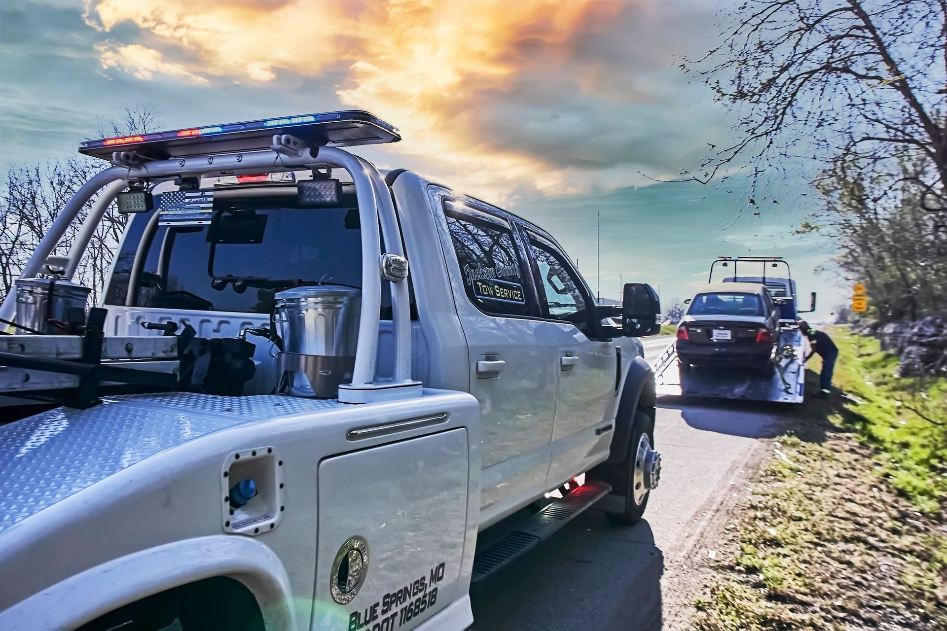 Tow truck with roof racks parked on a sunlit street, with another truck ahead at sunset.