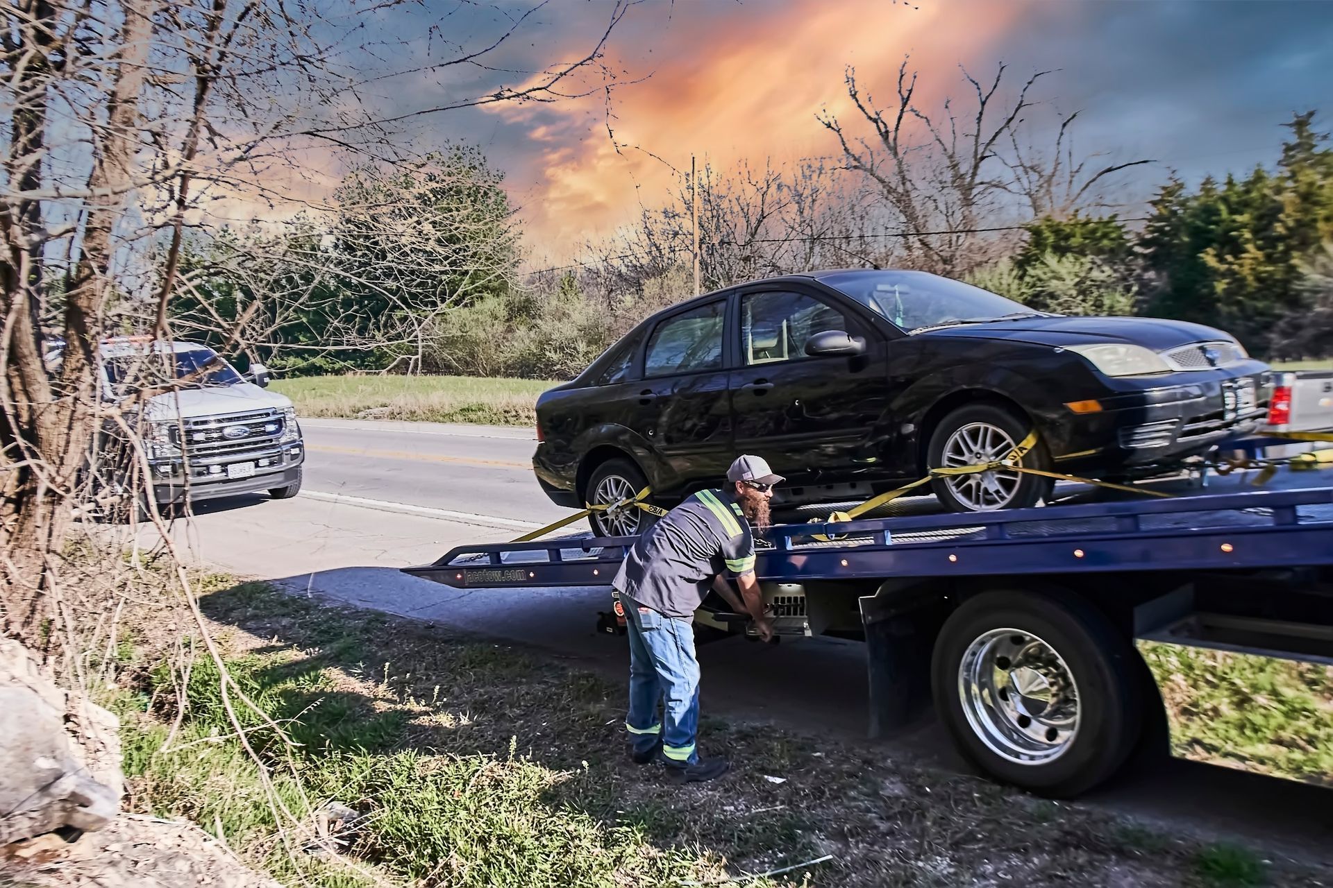 Tow truck operator loading a black sedan onto a flatbed at dusk after a roadside accident