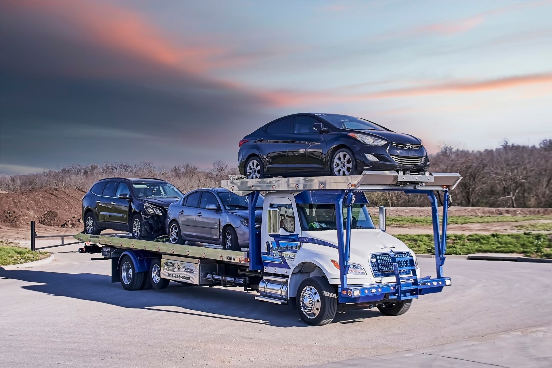 Blue car carrier truck hauling several new cars on a road at sunset