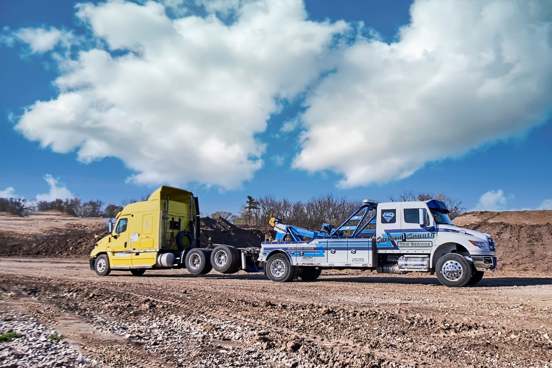 Yellow semi truck towing a blue-and-white flatbed trailer across a rocky desert under a cloudy sky