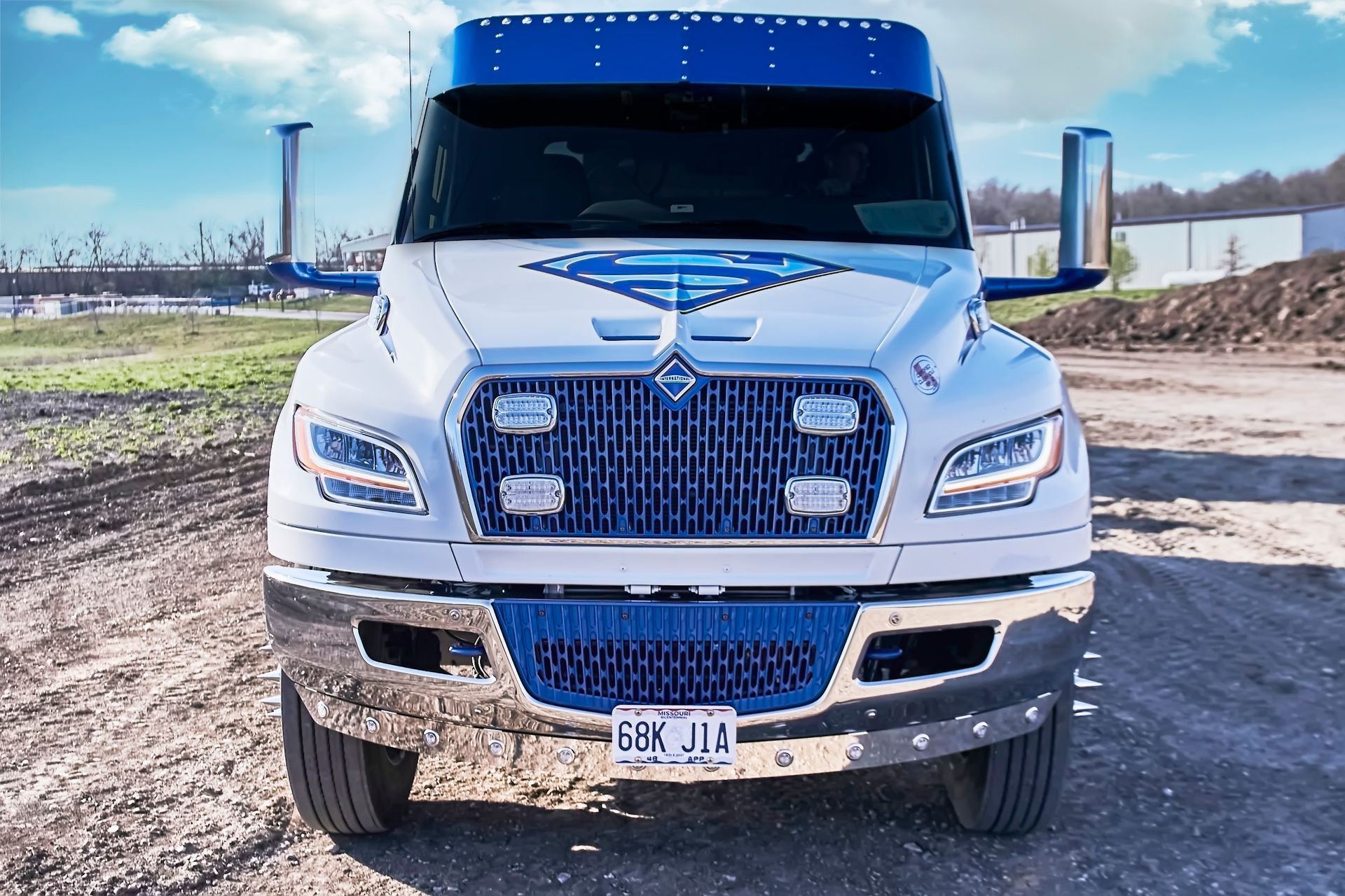 Front view of a white box truck on a dirt road, with a blue sky and industrial background.