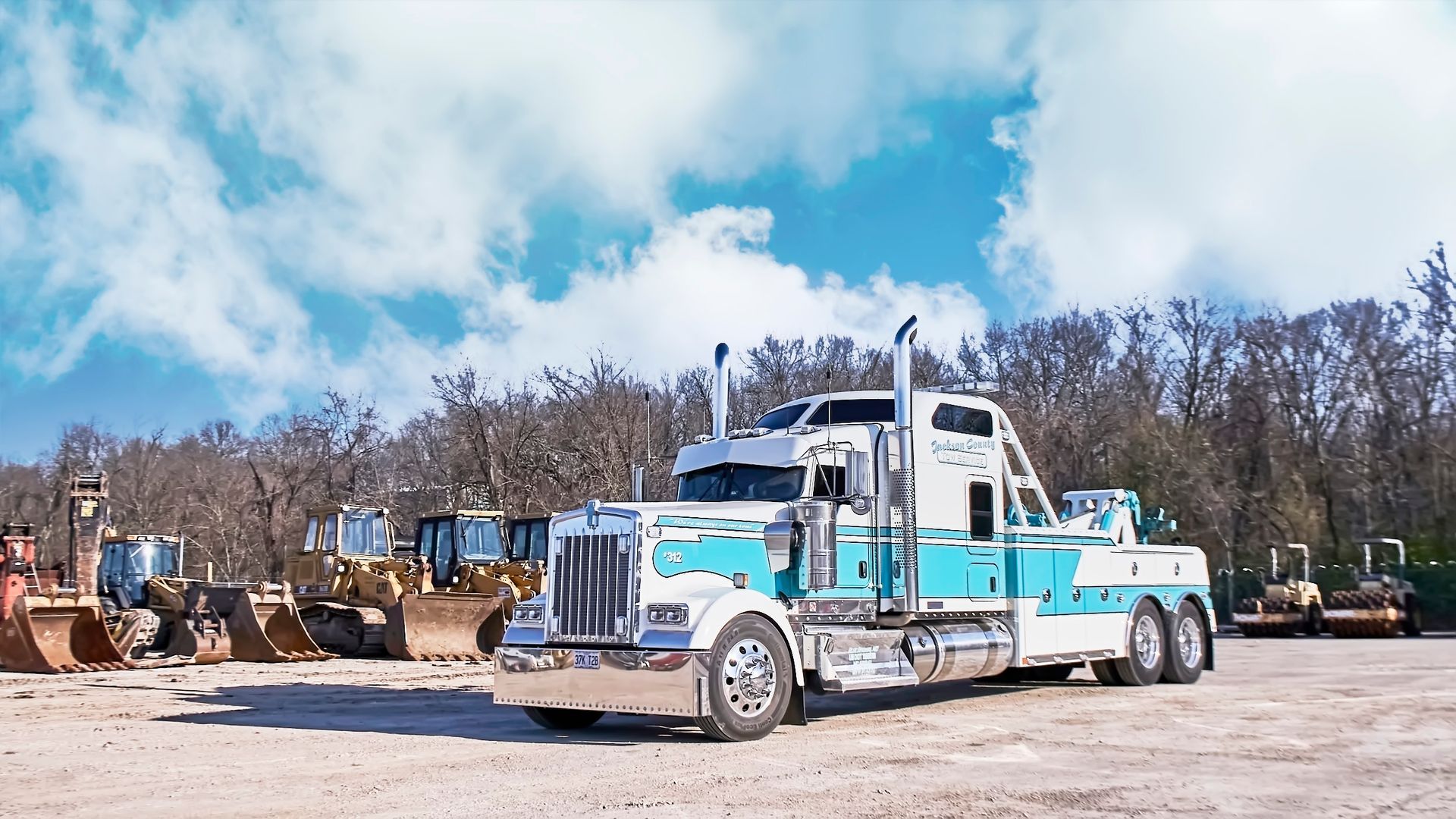 Blue-and-white tow truck parked in a gravel lot with heavy equipment and trees in the background