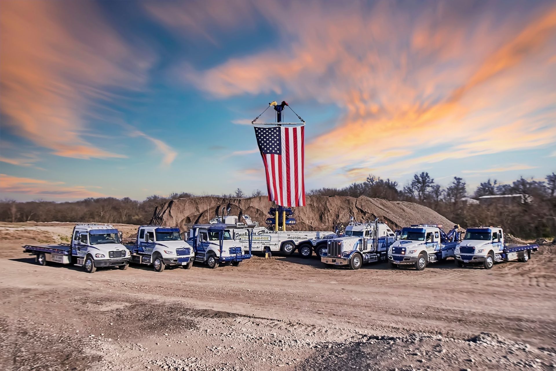 Line of white utility trucks beneath a giant American flag at sunset on a dusty lot.